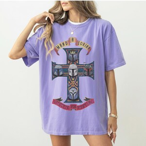 以下が含まれることがあります： ライトパープルの半袖Tシャツ。4つのマンダロリアン・ヘルメットが描かれた十字架のグラフィックが特徴です。十字架の上部のバナーには「MANDALORIAN」、下部のバナーには「APPETITE FOR HUNTING」という文字がプリントされています。
