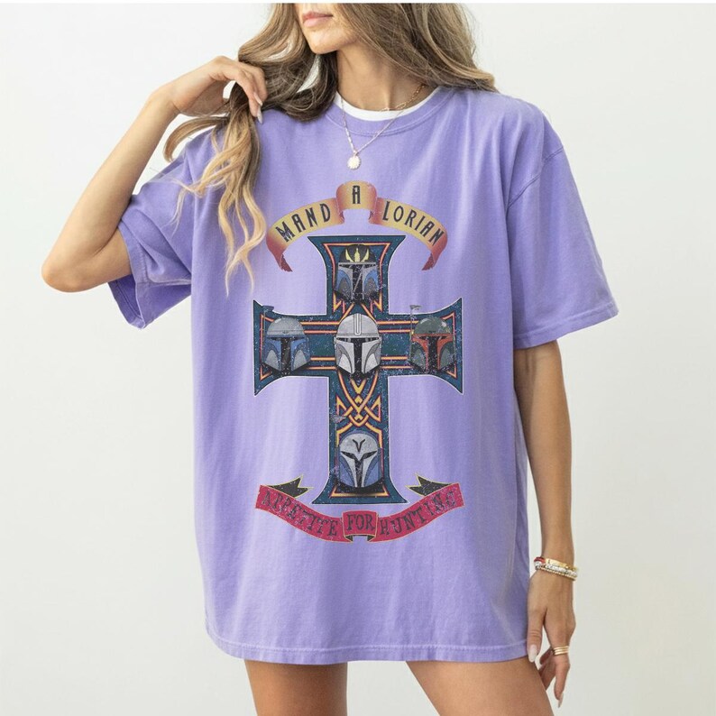 以下が含まれることがあります： ライトパープルの半袖Tシャツ。4つのマンダロリアン・ヘルメットが描かれた十字架のグラフィックが特徴です。十字架の上部のバナーには「MANDALORIAN」、下部のバナーには「APPETITE FOR HUNTING」という文字がプリントされています。