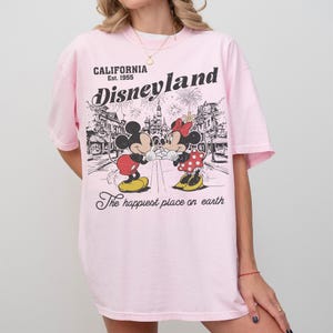 May include: A light pink t-shirt featuring a graphic of Mickey and Minnie Mouse in front of a Disneyland scene. The shirt has the words "CALIFORNIA Est. 1955 Disneyland" and "The happiest place on earth" printed on it.