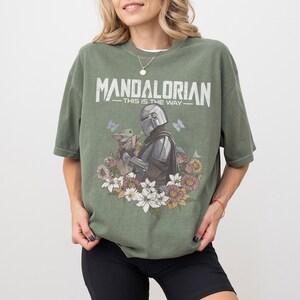 Op de afbeelding: Olijfgroen T-shirt met de tekst "MANDALORIAN - THIS IS THE WAY" in het wit. De afbeelding toont de Mandalorian en Grogu, omringd door bloemen en vlinders. Het T-shirt heeft een losse pasvorm.