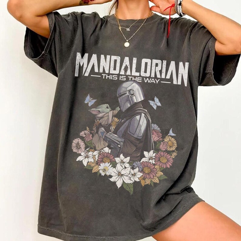Op de afbeelding: Donkergrijs t-shirt met de tekst "MANDALORIAN - THIS IS THE WAY". De afbeelding toont de Mandalorian en Grogu, omringd door bloemen en vlinders. Het shirt heeft een losse pasvorm.