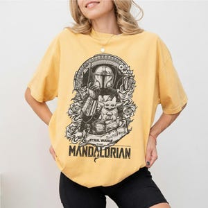 May include: Yellow t-shirt featuring a black and white graphic of The Mandalorian and Grogu from Star Wars. The design includes roses and the text "STAR WARS THE MANDALORIAN". The shirt has short sleeves and a crew neck.
