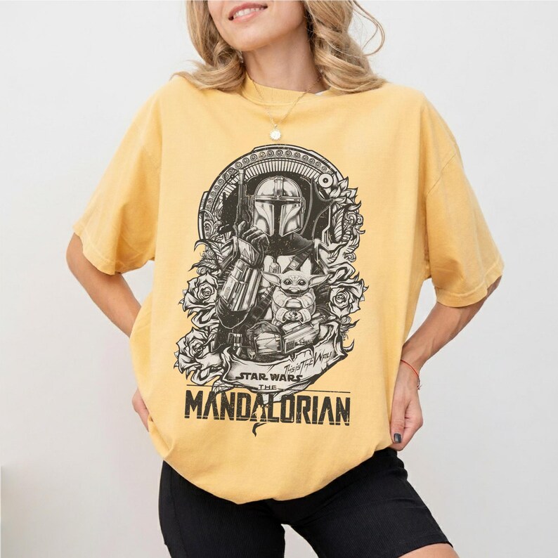 May include: Yellow t-shirt featuring a black and white graphic of The Mandalorian and Grogu from Star Wars. The design includes roses and the text "STAR WARS THE MANDALORIAN". The shirt has short sleeves and a crew neck.