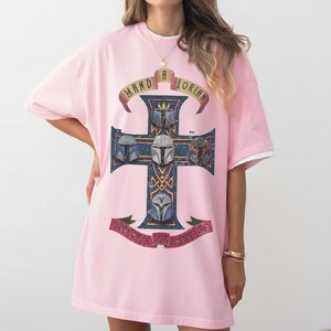 以下が含まれることがあります： ライトピンクの半袖Tシャツ。中央には4つのマンダロリアンのヘルメットが描かれたダークブルーの十字架のグラフィックがあります。十字架の上部のバナーには「MANDALORIAN」、下部のバナーには「BLESS THE HUNTERS」という文字がプリントされています。