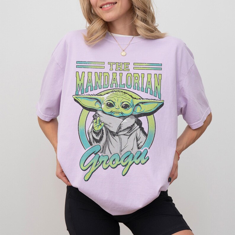 May include: A light purple t-shirt featuring a graphic of Grogu from The Mandalorian. The design includes the text "THE MANDALORIAN" and "Grogu" in green and blue, with an image of Grogu in the center.