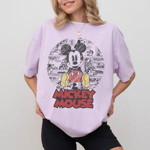 May include: A light purple t-shirt featuring a graphic of Mickey Mouse. The design includes a circular comic strip background and the words "Mickey Mouse" in red and yellow. The shirt has short sleeves and a relaxed fit.