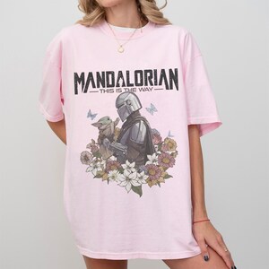 Op de afbeelding: Lichtroze T-shirt met de tekst "MANDALORIAN - THIS IS THE WAY" in zwart. De afbeelding toont de Mandalorian en Grogu, omringd door bloemen en vlinders. Het shirt heeft korte mouwen en een ronde hals.