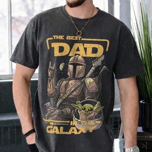 Pu&ograve; includere: T-shirt grigio scuro con una grafica che raffigura un personaggio Mandaloriano che suona la chitarra, con Grogu in una capsula. Il testo recita "THE BEST DAD IN THE GALAXY" in giallo. La maglietta ha un aspetto vintage.