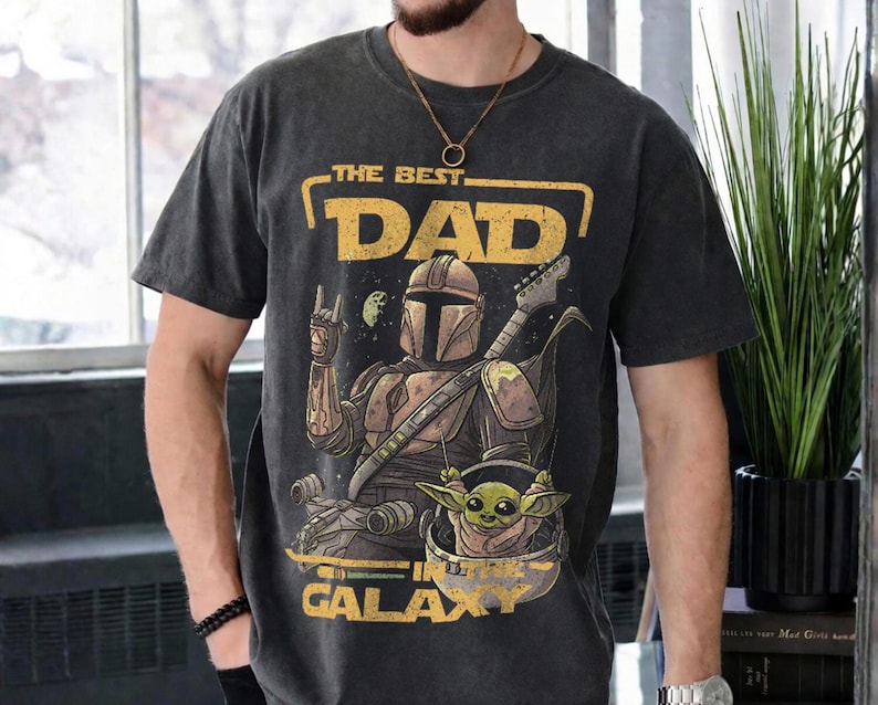 Pu&ograve; includere: T-shirt grigio scuro con una grafica che raffigura un personaggio Mandaloriano che suona la chitarra, con Grogu in una capsula. Il testo recita "THE BEST DAD IN THE GALAXY" in giallo. La maglietta ha un aspetto vintage.