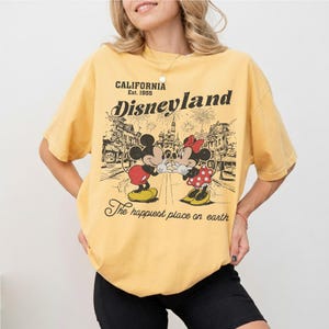 May include: Yellow t-shirt with a Disneyland graphic featuring Mickey and Minnie Mouse. The graphic includes the text "CALIFORNIA Est. 1955 Disneyland" and "The happiest place on earth."
