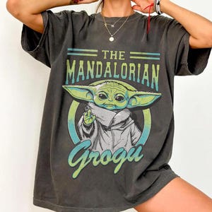 May include: Dark gray t-shirt featuring the text "THE MANDALORIAN" above an illustration of Grogu, also known as Baby Yoda. The Grogu graphic is in green, white, and blue. The word "Grogu" is written in a stylized font below the image.