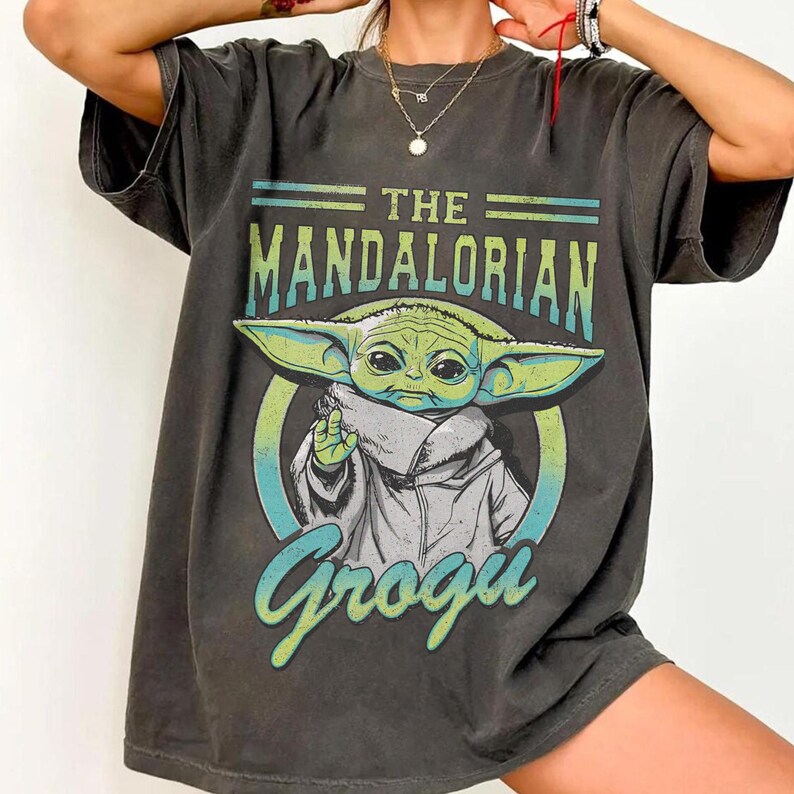 May include: Dark gray t-shirt featuring the text "THE MANDALORIAN" above an illustration of Grogu, also known as Baby Yoda. The Grogu graphic is in green, white, and blue. The word "Grogu" is written in a stylized font below the image.
