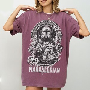 May include: A maroon t-shirt featuring a black and white graphic of The Mandalorian and Grogu from Star Wars. The design includes the text "STAR WARS THE MANDALORIAN" and floral accents. The shirt has short sleeves and a crew neck.