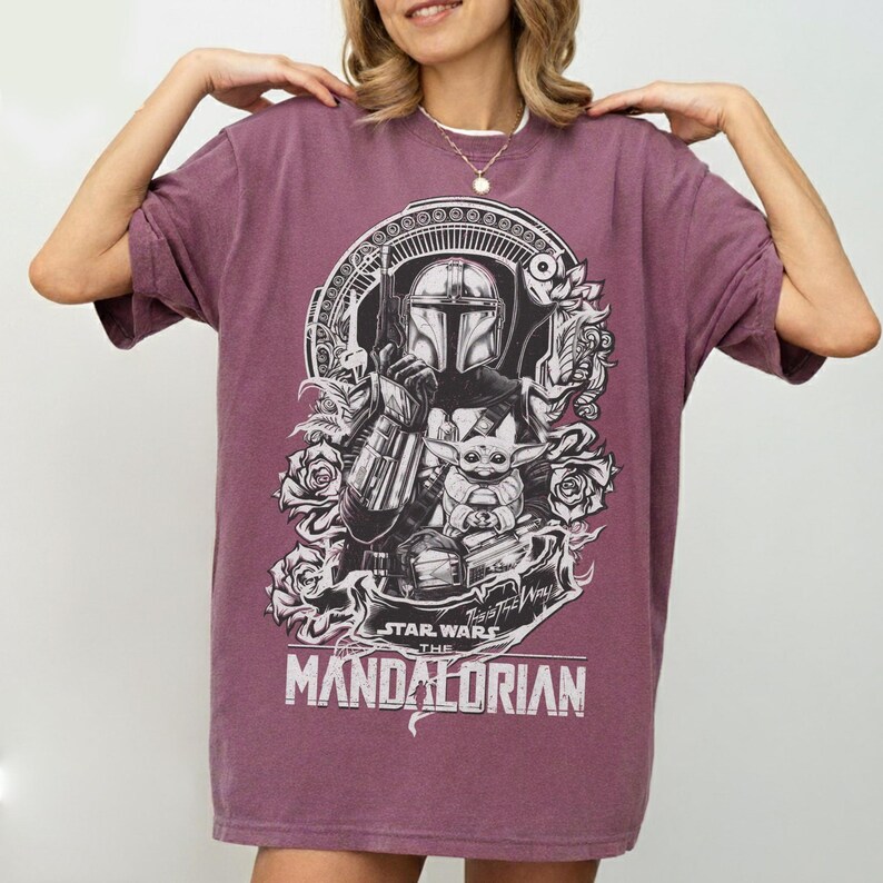 May include: A maroon t-shirt featuring a black and white graphic of The Mandalorian and Grogu from Star Wars. The design includes the text "STAR WARS THE MANDALORIAN" and floral accents. The shirt has short sleeves and a crew neck.