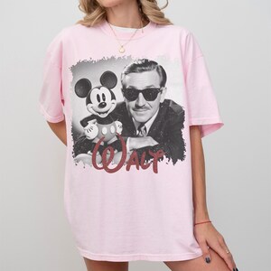 K&ouml;nnte beinhalten: Hellrosa T-Shirt mit einem Schwarz-Wei&szlig;-Bild von Walt Disney und Mickey Mouse. Das Wort "WALT" ist in roter Schrift darunter gedruckt. Das Shirt hat kurze &Auml;rmel und einen Rundhalsausschnitt.