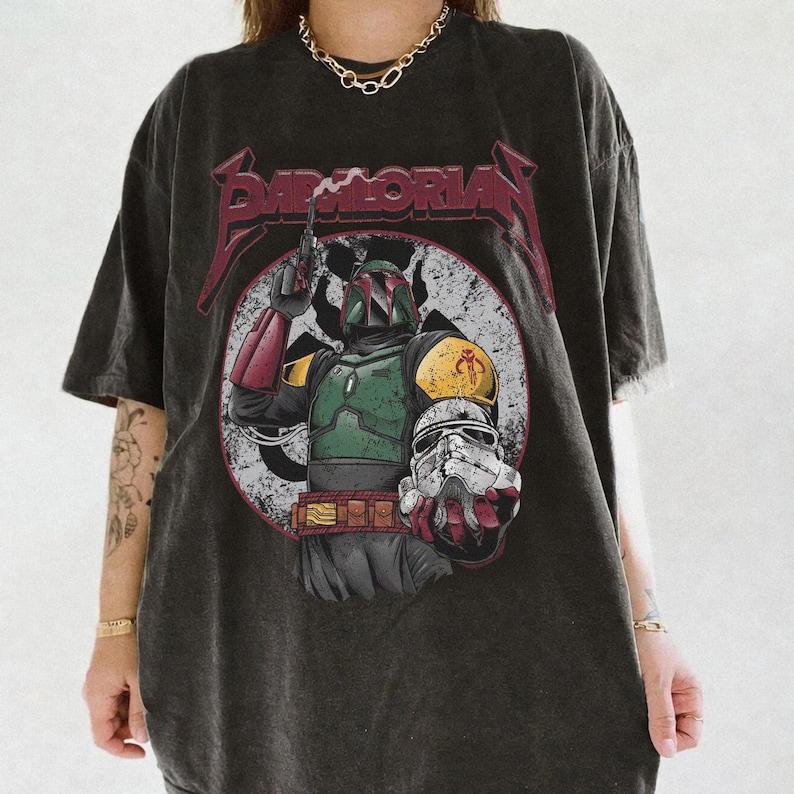 May include: Dark gray t-shirt featuring a graphic of Boba Fett holding a blaster and a Stormtrooper helmet. The design is set within a white circle with a distressed look, and the word "Mandalorian" is printed above the image in red. The shirt has short sleeves.