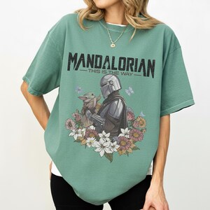 Op de afbeelding: Groen t-shirt met een afbeelding van The Mandalorian en Grogu omringd door bloemen en vlinders. De tekst "MANDALORIAN - THIS IS THE WAY" staat in het zwart bovenaan.
