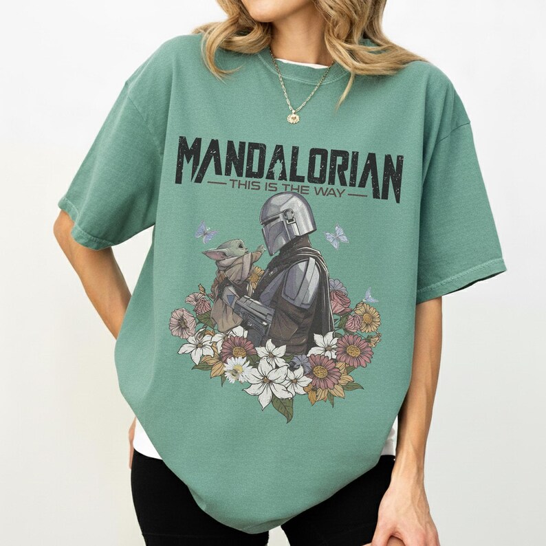 Op de afbeelding: Groen t-shirt met een afbeelding van The Mandalorian en Grogu omringd door bloemen en vlinders. De tekst "MANDALORIAN - THIS IS THE WAY" staat in het zwart bovenaan.