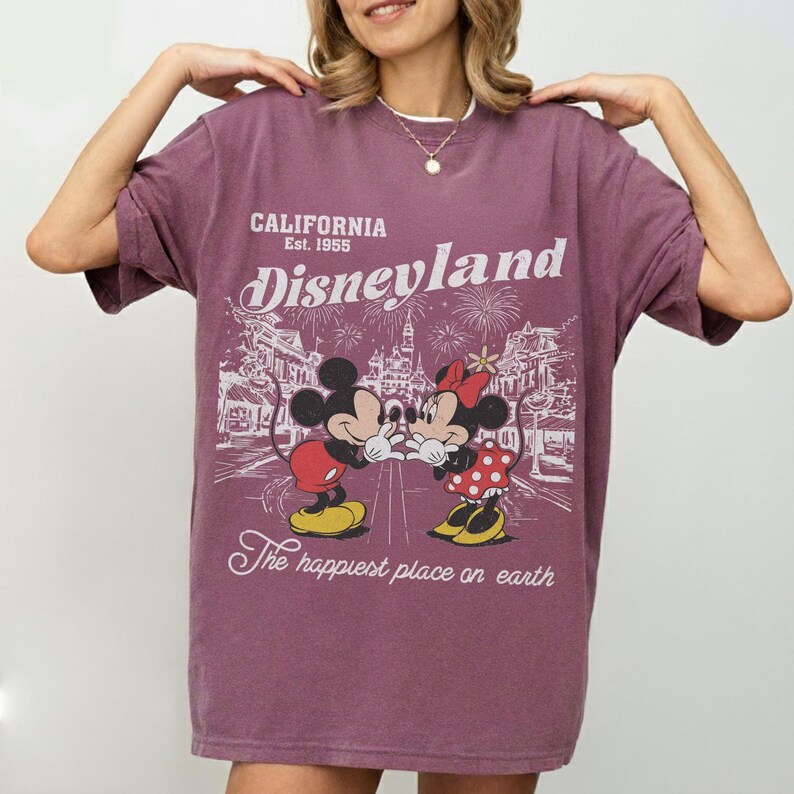 May include: A maroon Disneyland t-shirt featuring Mickey and Minnie Mouse kissing. The shirt has the text "CALIFORNIA Est. 1955 Disneyland" and "The happiest place on earth" in white. The graphic shows a street scene with fireworks.