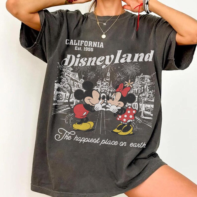 May include: Dark gray t-shirt featuring Mickey and Minnie Mouse in front of a Disneyland scene with fireworks. The shirt has the text "CALIFORNIA Est. 1955 Disneyland" and "The happiest place on earth".
