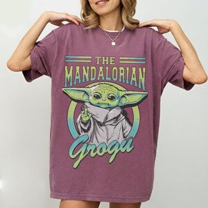 May include: A maroon t-shirt featuring a graphic of Grogu from The Mandalorian. The design includes the text "THE MANDALORIAN" above an image of Grogu, and the name "Grogu" below. The Grogu image is green and white.