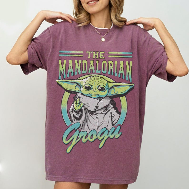 May include: A maroon t-shirt featuring a graphic of Grogu from The Mandalorian. The design includes the text "THE MANDALORIAN" above an image of Grogu, and the name "Grogu" below. The Grogu image is green and white.