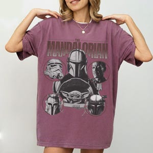 Pu&ograve; includere: T-shirt color amaranto con un design grafico della serie "The Mandalorian". Il design include il titolo dello show e immagini di personaggi come il Mandaloriano, uno Stormtrooper e Grogu. La maglietta ha maniche corte e girocollo.