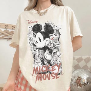 May include: Cream-colored t-shirt featuring a black and white graphic of Mickey Mouse with the word "Disney" above and "Mickey Mouse" in red below. The design has a street art style.