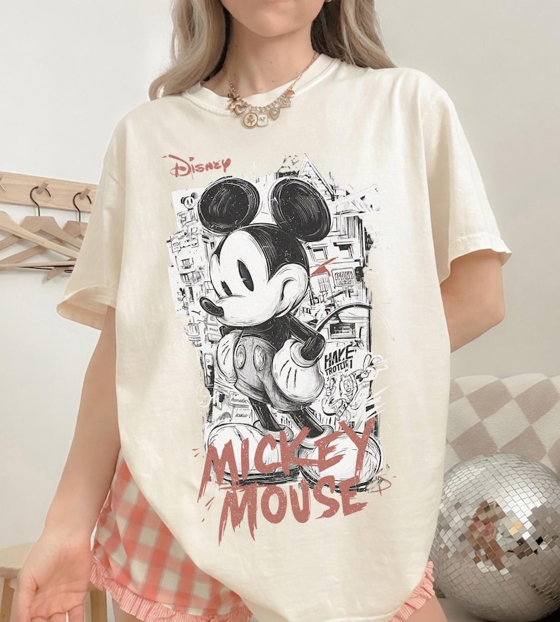 May include: Cream-colored t-shirt featuring a black and white graphic of Mickey Mouse with the word "Disney" above and "Mickey Mouse" in red below. The design has a street art style.
