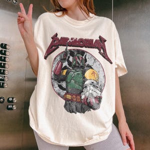 May include: Cream-colored t-shirt featuring a graphic of Boba Fett and a Stormtrooper helmet within a circular design. The shirt has the text "BAD MORIA" in a maroon, distressed font. The design is vintage-inspired.
