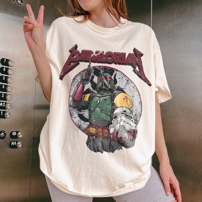 May include: Cream-colored t-shirt featuring a graphic of Boba Fett and a Stormtrooper helmet within a circular design. The shirt has the text "BAD MORIA" in a maroon, distressed font. The design is vintage-inspired.