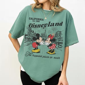 May include: Green t-shirt featuring a graphic of Mickey and Minnie Mouse in front of Disneyland. The shirt has the text "CALIFORNIA Est. 1955 Disneyland" and "The happiest place on earth."