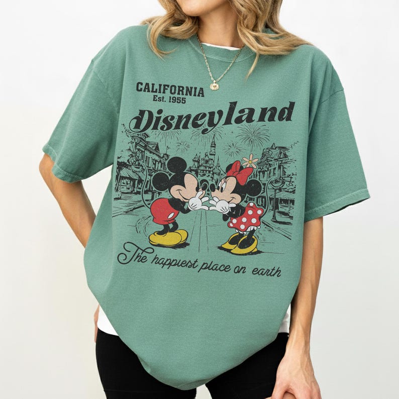 May include: Green t-shirt featuring a graphic of Mickey and Minnie Mouse in front of Disneyland. The shirt has the text "CALIFORNIA Est. 1955 Disneyland" and "The happiest place on earth."