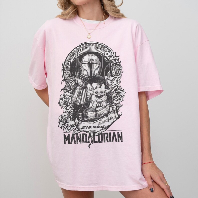 May include: A light pink t-shirt featuring a black and white graphic of The Mandalorian and Grogu from Star Wars. The design includes roses and the text "STAR WARS THE MANDALORIAN".