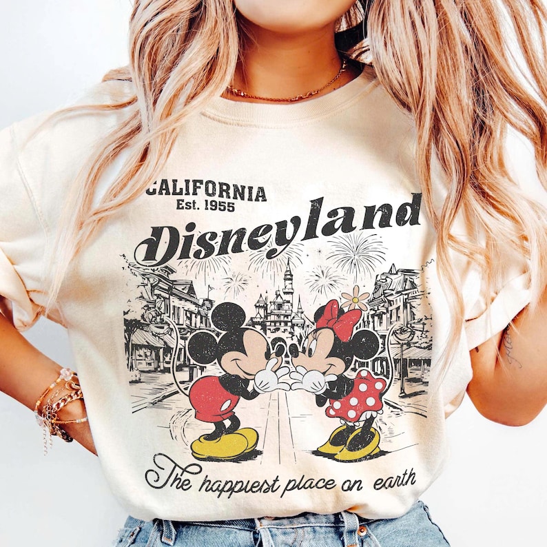 May include: Cream-colored Disneyland sweatshirt featuring Mickey and Minnie Mouse in a vintage design. The graphic includes the words "California Est. 1955" and "Disneyland" with fireworks and a cityscape. The phrase "The happiest place on earth" is also included.