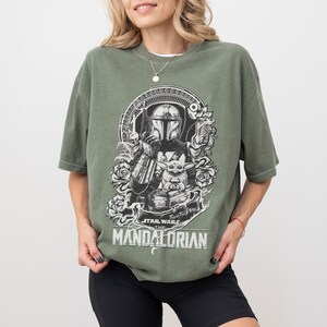 May include: Olive green t-shirt featuring a black and white graphic of The Mandalorian and Grogu. The design includes roses and the text "Star Wars The Mandalorian". The shirt has short sleeves and a relaxed fit.