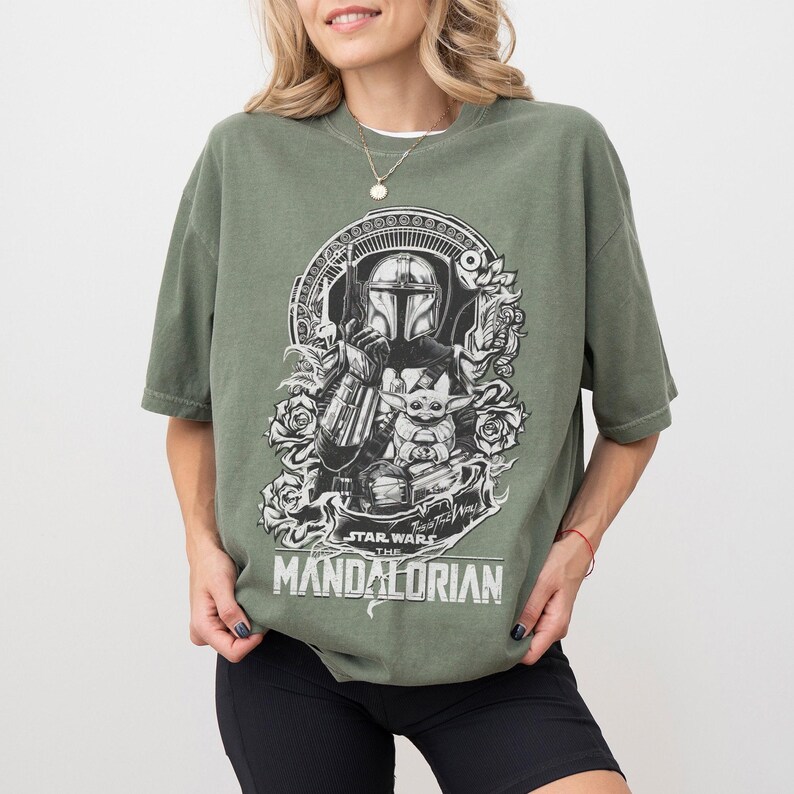 May include: Olive green t-shirt featuring a black and white graphic of The Mandalorian and Grogu. The design includes roses and the text "Star Wars The Mandalorian". The shirt has short sleeves and a relaxed fit.