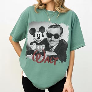 K&ouml;nnte beinhalten: Gr&uuml;nes T-Shirt mit einem Schwarz-Wei&szlig;-Bild von Mickey Mouse und Walt Disney. Das Wort "WALT" ist in roter Schrift geschrieben. Das T-Shirt hat kurze &Auml;rmel und einen Rundhalsausschnitt. Das T-Shirt hat eine lockere Passform.