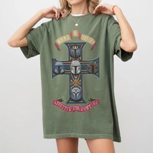 以下が含まれることがあります： オリーブグリーンの半袖Tシャツ。ゴールドの縁取りが施されたダークブルーの十字架のグラフィックが特徴です。十字架には5つのヘルメットが描かれ、上部には「MAND A LORIAN」、下部の赤いバナーには「APPETITE FOR HUNTING」という文字がプリントされています。