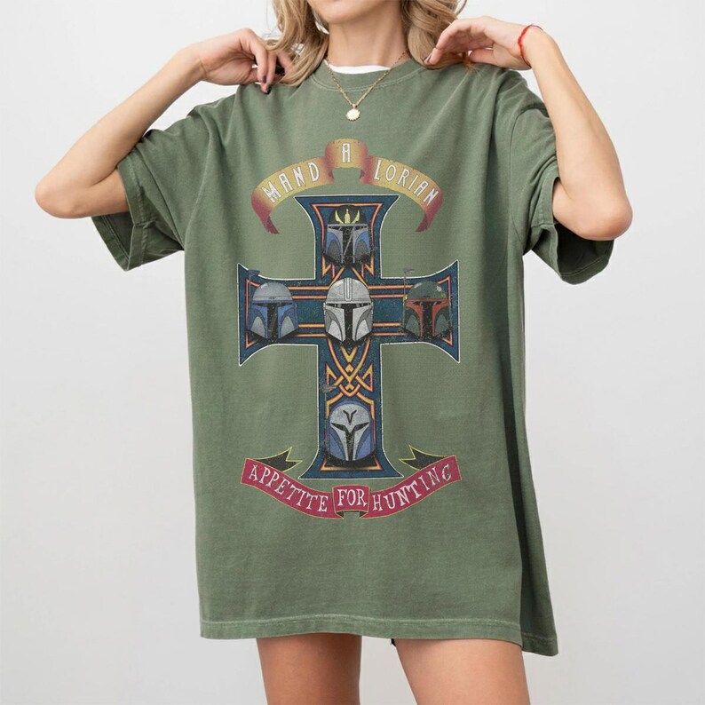 以下が含まれることがあります： オリーブグリーンの半袖Tシャツ。ゴールドの縁取りが施されたダークブルーの十字架のグラフィックが特徴です。十字架には5つのヘルメットが描かれ、上部には「MAND A LORIAN」、下部の赤いバナーには「APPETITE FOR HUNTING」という文字がプリントされています。