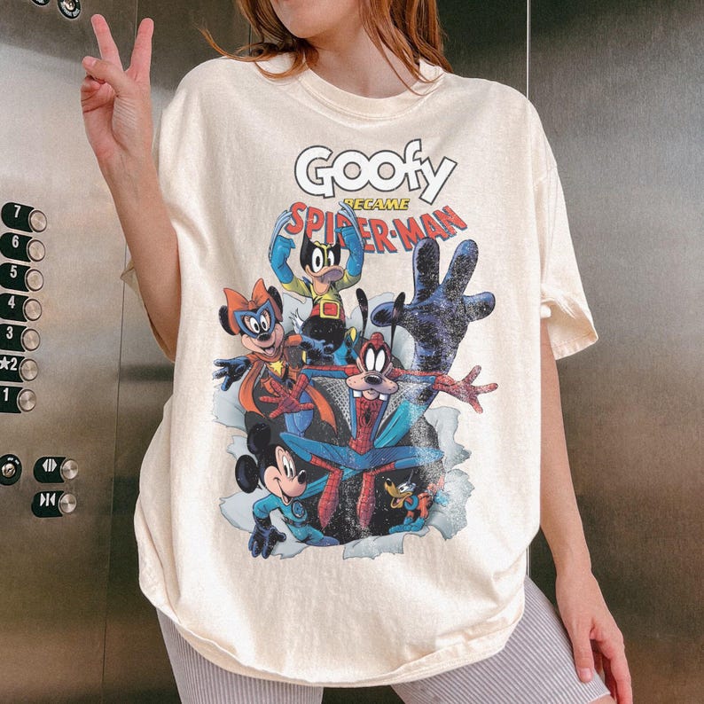 Op de afbeelding: Cr&egrave;mekleurig T-shirt met een afbeelding van Goofy als Spider-Man en andere Disney-personages. Het ontwerp bevat de tekst "Goofy Became Spider-Man". Het shirt heeft een ronde hals en korte mouwen.