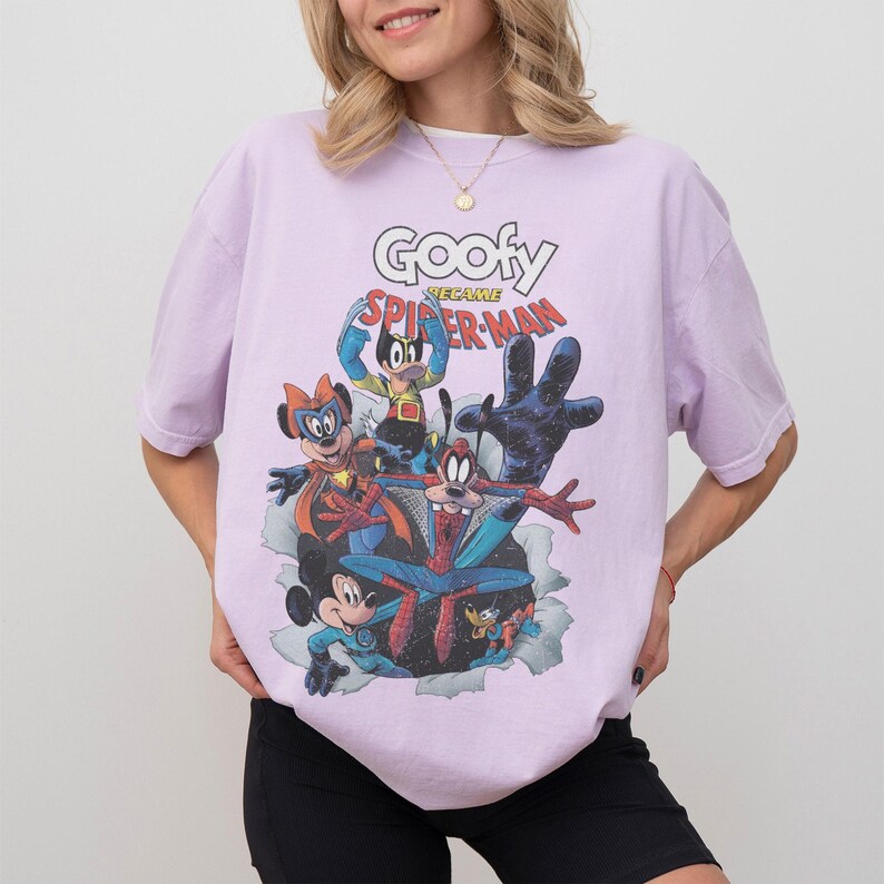 Op de afbeelding: Lichtpaars T-shirt met een stripboekafbeelding van Disney-personages als superhelden. De tekst "Goofy Became Spider-Man" staat boven de personages. Het shirt heeft korte mouwen.