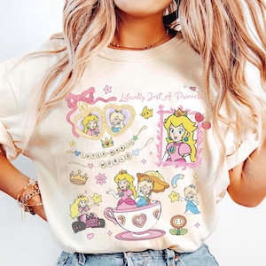 May include: Cream-colored sweatshirt featuring a colorful graphic of Princess Peach from Super Mario World. The design includes various illustrations of the princess, text that reads "Literally Just A Princess", and other themed elements.