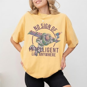 Op de afbeelding: Mosterdgeel T-shirt met een afbeelding van Buzz Lightyear en de tekst "NO SIGN OF INTELLIGENT LIFE ANYWHERE". De afbeelding toont Buzz in zijn ruimtepak met uitgestrekte vleugels en een laserpistool.