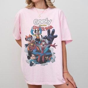 Op de afbeelding: Lichtroze T-shirt met een cartoonafbeelding van Disney-personages als superhelden. Het ontwerp bevat de tekst "Goofy Became Spider-Man". Het shirt heeft korte mouwen en een ronde hals.
