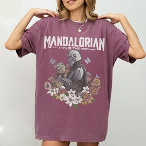 Op de afbeelding: Bordeauxrood T-shirt met het woord "MANDALORIAN" en de zin "THIS IS THE WAY". Het shirt heeft een illustratie van een personage en Grogu, omringd door bloemen en vlinders.