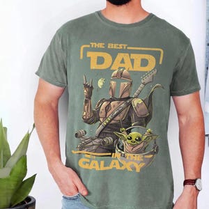 Pu&ograve; includere: T-shirt verde con una grafica raffigurante un personaggio in stile Mandaloriano che suona la chitarra, con Grogu in una capsula. Il testo recita "THE BEST DAD IN THE GALAXY" in giallo. La maglietta &egrave; indossata da una persona.
