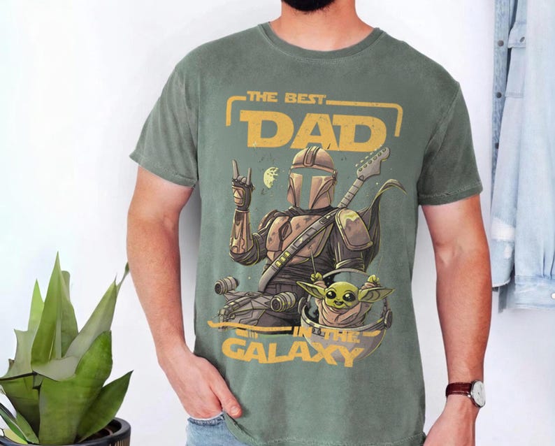 Pu&ograve; includere: T-shirt verde con una grafica raffigurante un personaggio in stile Mandaloriano che suona la chitarra, con Grogu in una capsula. Il testo recita "THE BEST DAD IN THE GALAXY" in giallo. La maglietta &egrave; indossata da una persona.