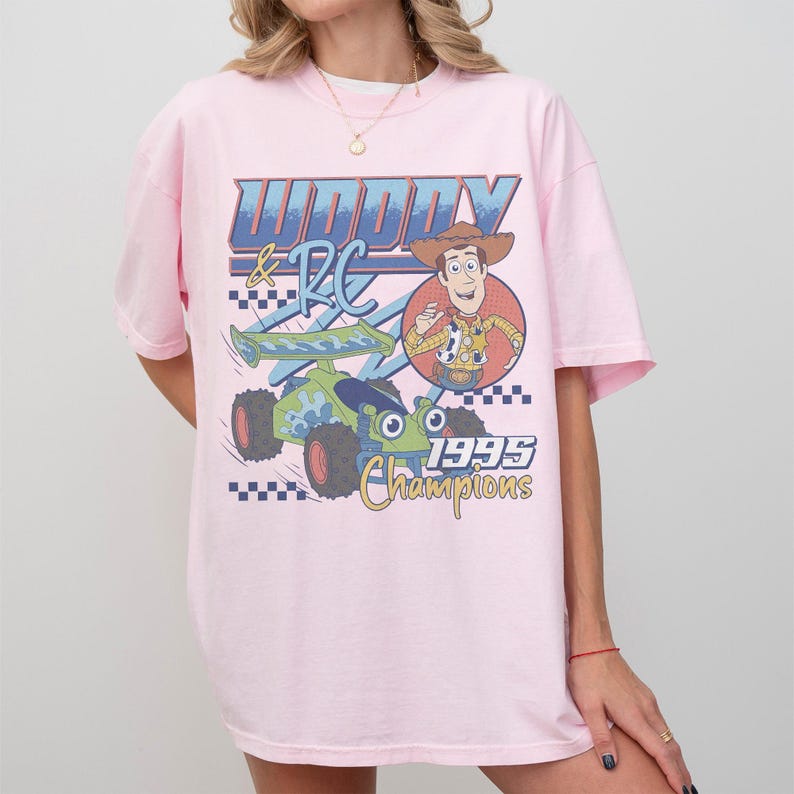 Puede incluir: Camiseta rosa claro con estampado gr&aacute;fico. El dise&ntilde;o presenta el texto "Woody & RC 1995 Champions" y una imagen de Woody y un coche de carreras verde y azul.