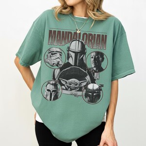 Pu&ograve; includere: T-shirt verde con un design grafico con personaggi di The Mandalorian. Il design include il testo "MANDALORIAN" e immagini di un casco da Stormtrooper, The Mandalorian, Grogu e altri personaggi.